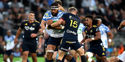 Super Rugby Pacific action from Round 2 as players from the Blues and Highlanders battle for possession in a high-intensity match. A Highlanders fullback attempts a tackle on a Blues forward, with teammates in pursuit under the stadium lights.