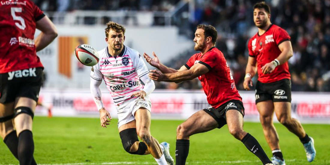 A Stade Français player offloads the ball under pressure from a Toulon defender during their Top 14 Round 17 clash. Toulon players close in as the intensity of the match unfolds on the field.