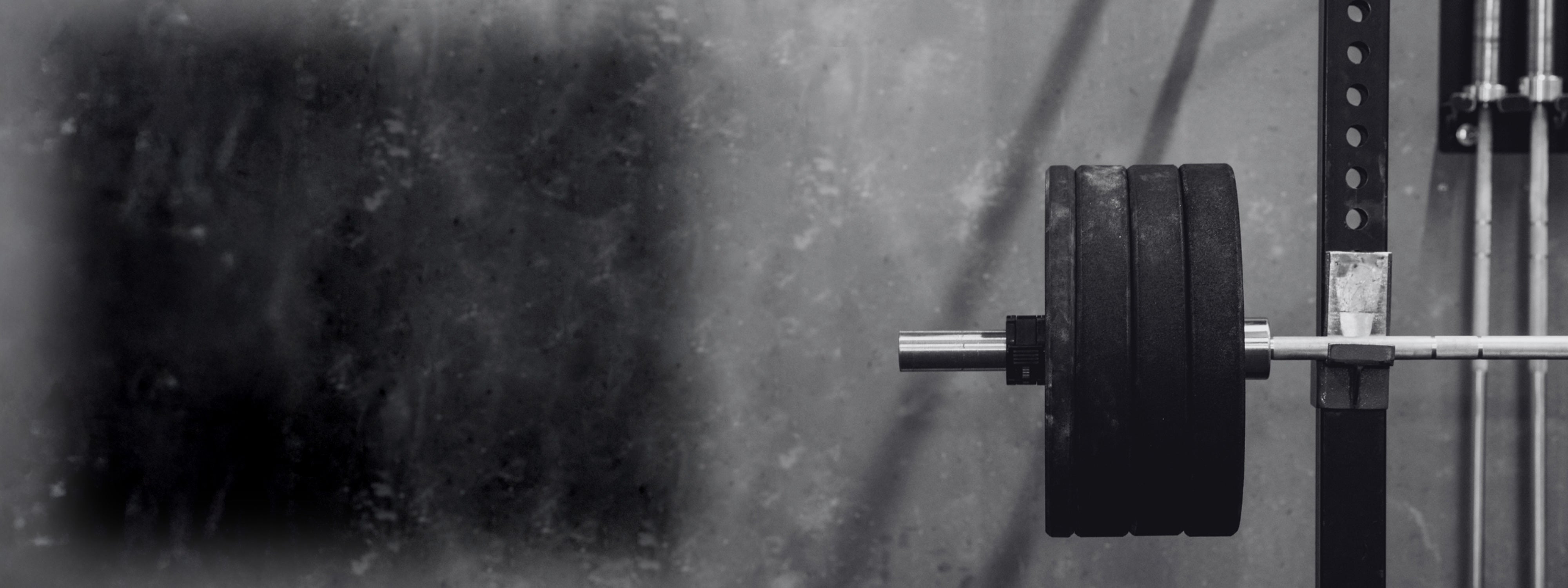 Black and white background image of a barbell with weight plates on a squat rack, set against a rugged gym wall. Used as the hero banner image for the PEAK RUGBY Training Page.